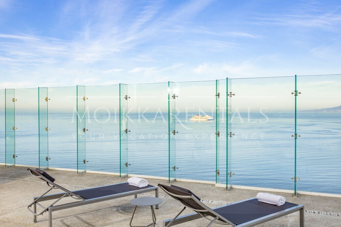 A glass-enclosed balcony with two sun loungers and a small table, overlooking a calm sea with a ship in the distance.