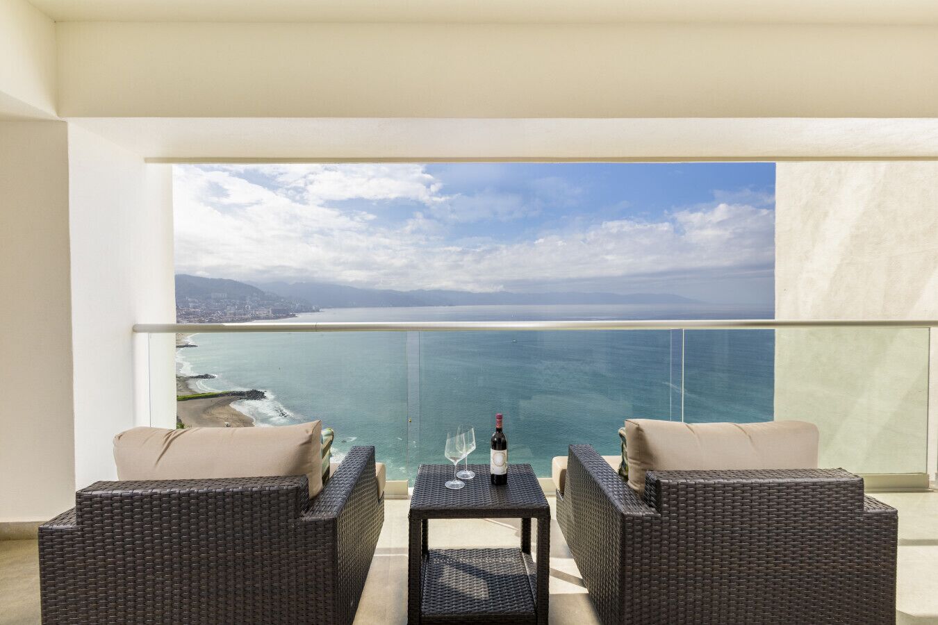 A balcony with wicker furniture, a glass table, and a bottle of wine.