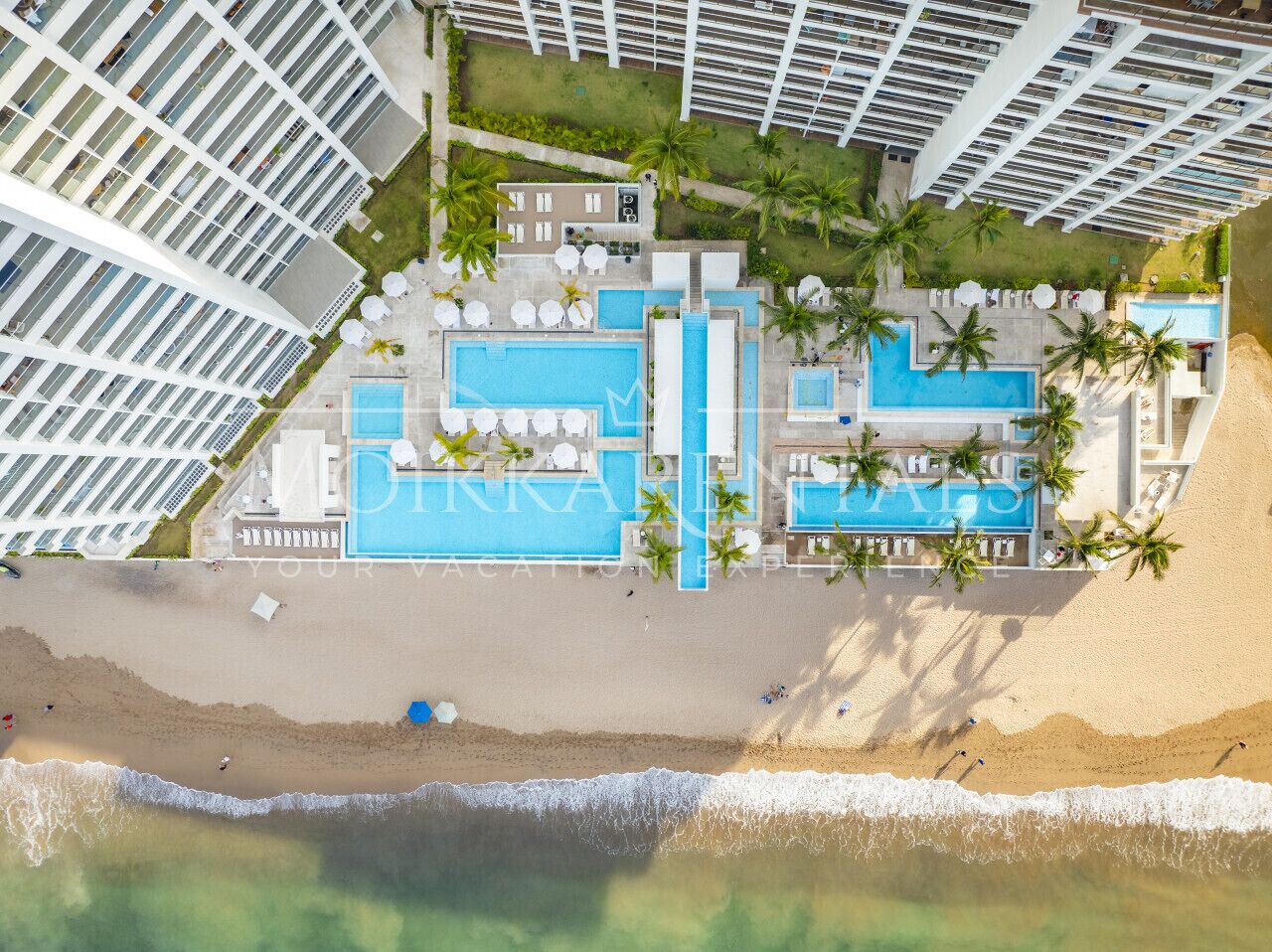 Vista aérea de un complejo turístico con varias piscinas, una playa y edificios altos.