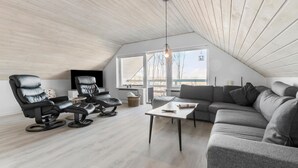 A modern loft bedroom with a sloped ceiling, a large window, a sofa, a recliner, and a flat-screen TV.