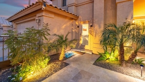 A building with a tiled roof, a well-lit entrance, and a landscaped garden with palm trees. - Private Guest Suite - Pool, Hot Tub, Cabana, Courtyard & Putting Green (Peoria)