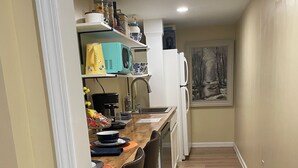 Een smalle keuken met een houten aanrecht, een spoelbak en een koelkast. Er hangt een schilderij aan de muur en een raam met uitzicht op bomen.