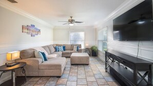 A modern living room with a sectional sofa, a flat-screen TV, and a ceiling fan. - Pool + Tennis/B-Ball Courts + Game Room + Mins to Disney! (Kissimmee)