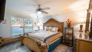 A bedroom with a wooden bed, a ceiling fan, a window with blinds, a bedside table with a lamp, and a decorative mirror. - Pool + Tennis/B-Ball Courts + Game Room + Mins to Disney! (Kissimmee)