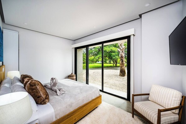 A modern bedroom with a large bed, a chair, and a view of a garden through floor-to-ceiling windows.