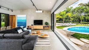 A modern living room with a large TV, grey sofa, wooden coffee table, and a view of a swimming pool and greenery. - Luxury Modern Escape in Hacienda Pinilla: Beach Club Access & Endless Adventure! (Pinilla)