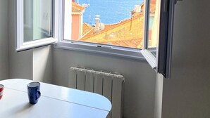 A room with a large window overlooking a coastal view, a white table with two mugs, and a radiator. - PEARL AP4444 By Riviera Holiday Homes (Villefranche-sur-Mer)