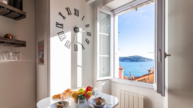 A dining area with a table set for two, a large wall clock, and a window with a view of the sea. - PEARL AP4444 By Riviera Holiday Homes (Villefranche-sur-Mer)