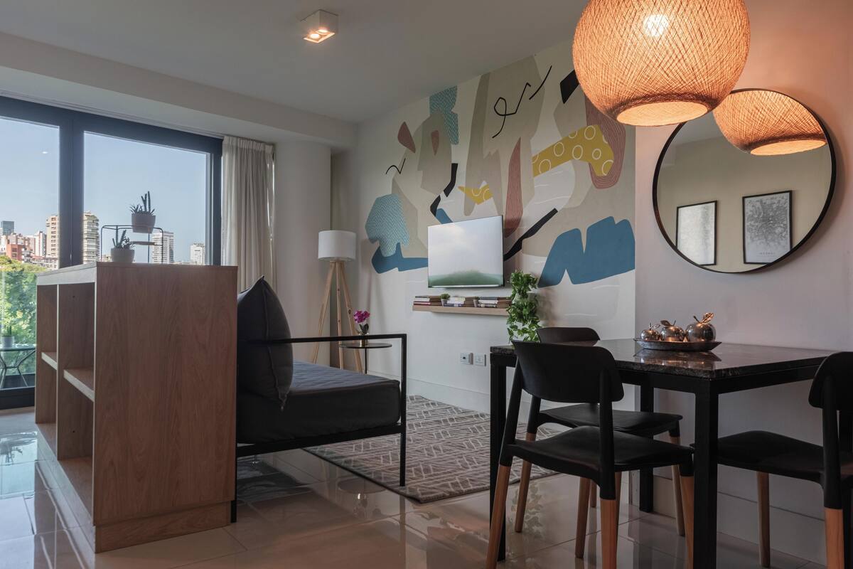 A modern living room with a sofa, dining table, chairs, a coffee table, a TV, a large window, and a city view.