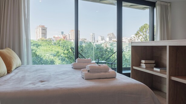 A hotel room with a large bed, two towels, a wooden shelf with books, and a view of a cityscape through large windows. - MOT MANSION BOTANICAL (Buenos Aires)
