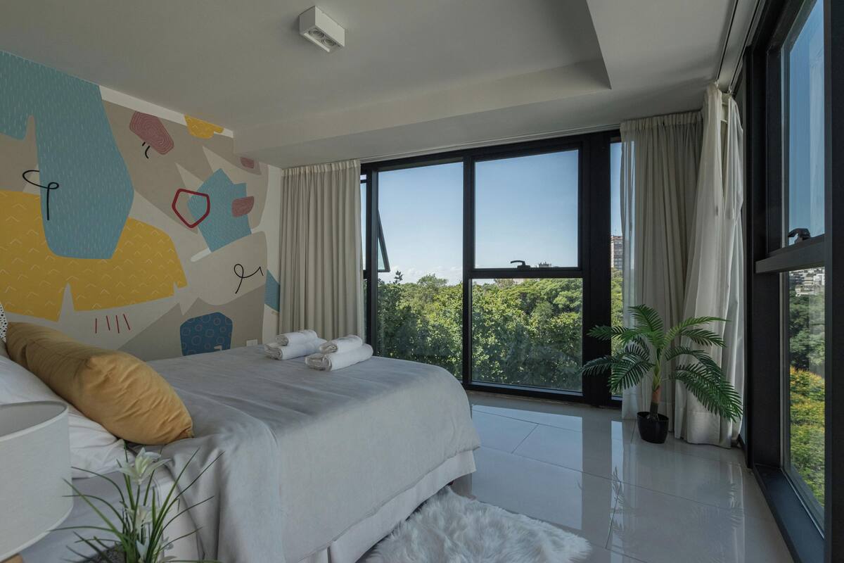 A modern bedroom with a large bed, a view of greenery outside, and a ceiling-mounted air conditioning unit.
