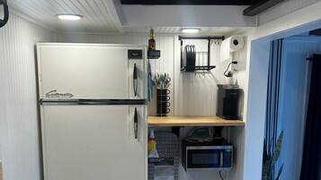 A compact kitchen with a white refrigerator, a microwave, and a kettle. There is a small shelf with kitchen utensils and a kettle. The kitchen has a wooden floor and a patterned rug.
