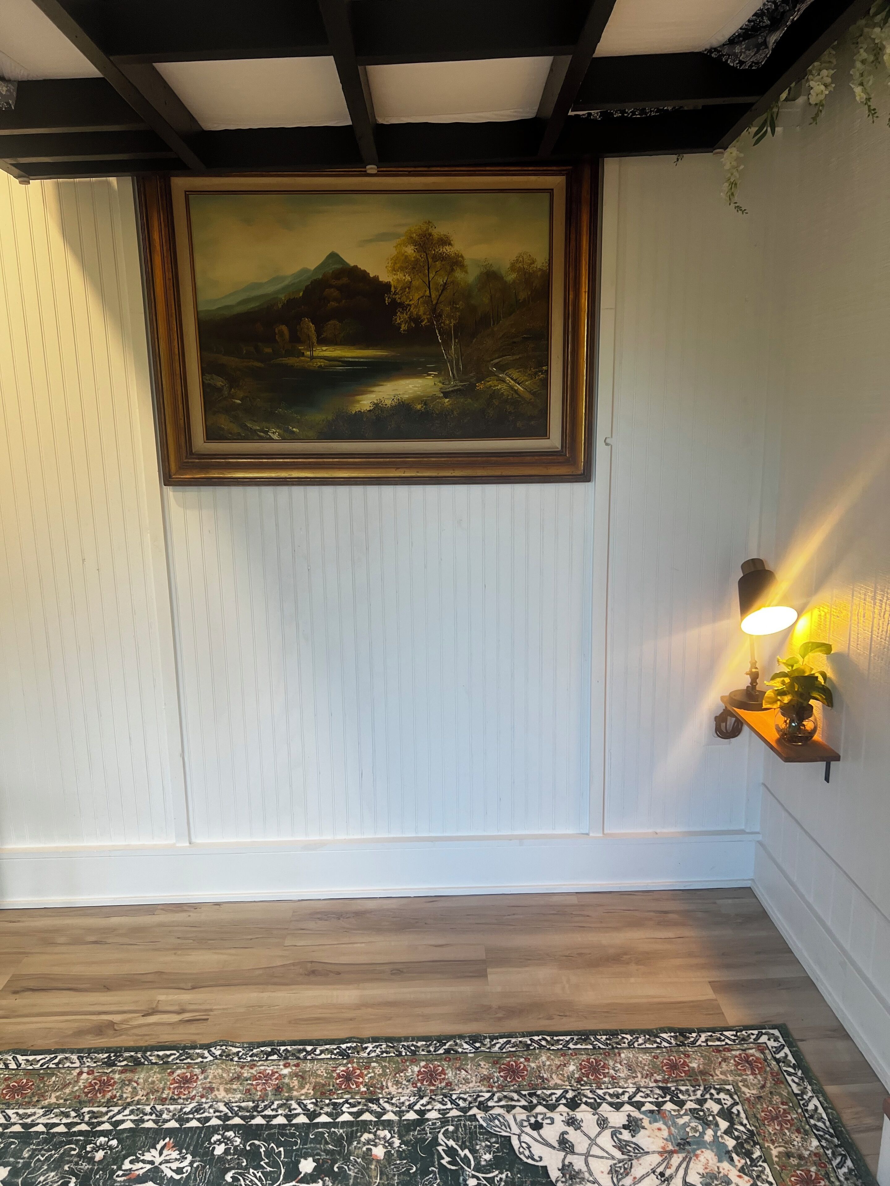 Une pièce avec un tableau représentant un paysage, encadré, un plancher en bois et un tapis à motifs.