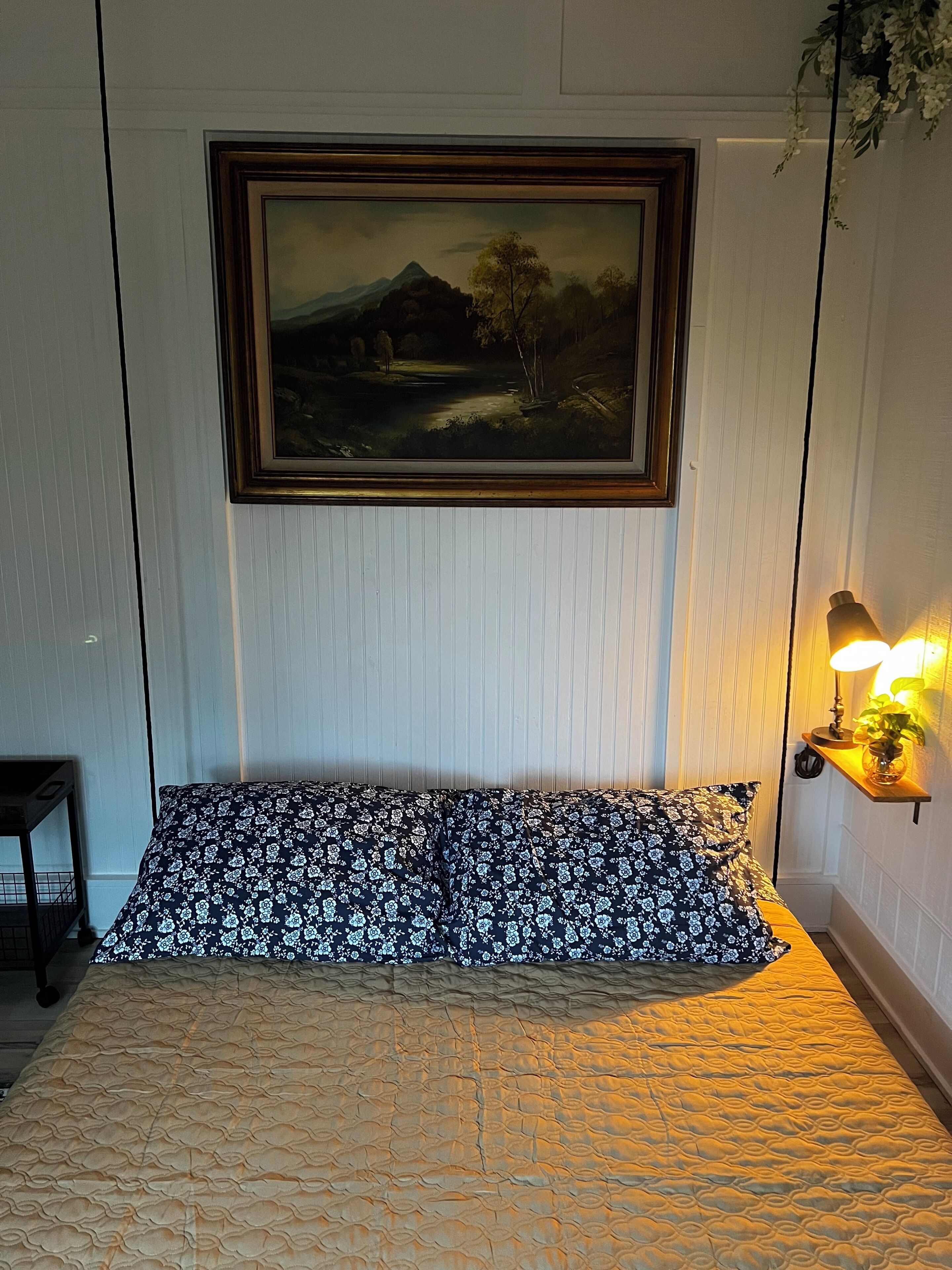 Un dormitorio con cama, un cuadro enmarco de paisaje, una lámpara de noche y una mesita con una planta.