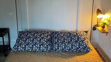 Un dormitorio con cama, un cuadro enmarco de paisaje, una lámpara de noche y una mesita con una planta.