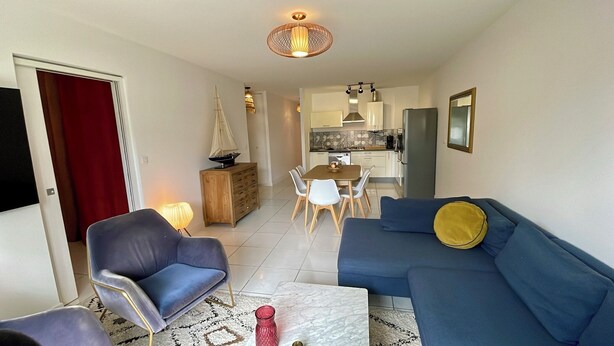 Una sala de estar moderna con un sofá azul, un comedor con sillas blancas y una cocina al fondo.