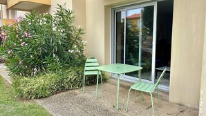 Un espacio al aire libre con mesa y dos sillas, un pequeño jardín con diversas plantas, y un edificio con un amplio ventanal.