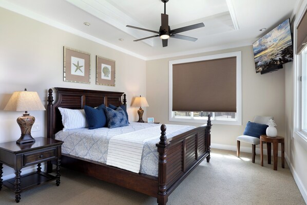 A bedroom with a wooden bed, two bedside tables with lamps, a ceiling fan, a large window with blinds, and artwork on the walls. - Mauna Kea - Waiulaula Condo - Stunning Sunset and Sunrise Views - Close to Pool (Kamuela)