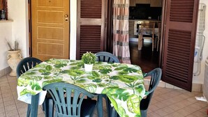 A dining area with a table covered in a tropical leaf tablecloth, surrounded by plastic chairs. A wooden door with a number plate is visible to the left. - Apartment 5 min from Airport, Self Check-in h24, Private Garden, Wi-Fi, A/C, TV (Rome Fiumicino)