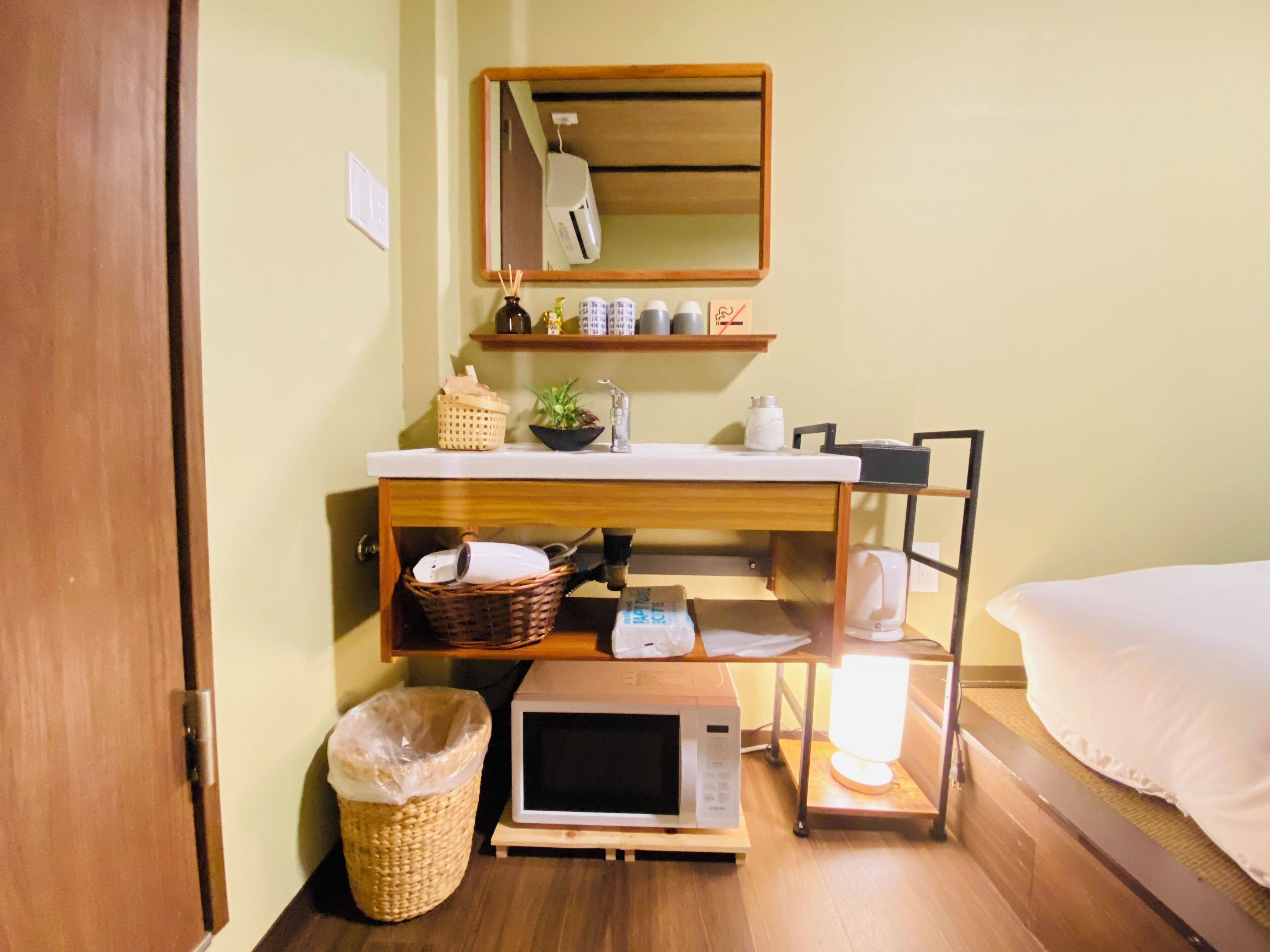 A compact hotel room with a wooden bed, a small bedside table with a lamp, a microwave, a trash bin, a wicker basket, and a shelf with toiletries.