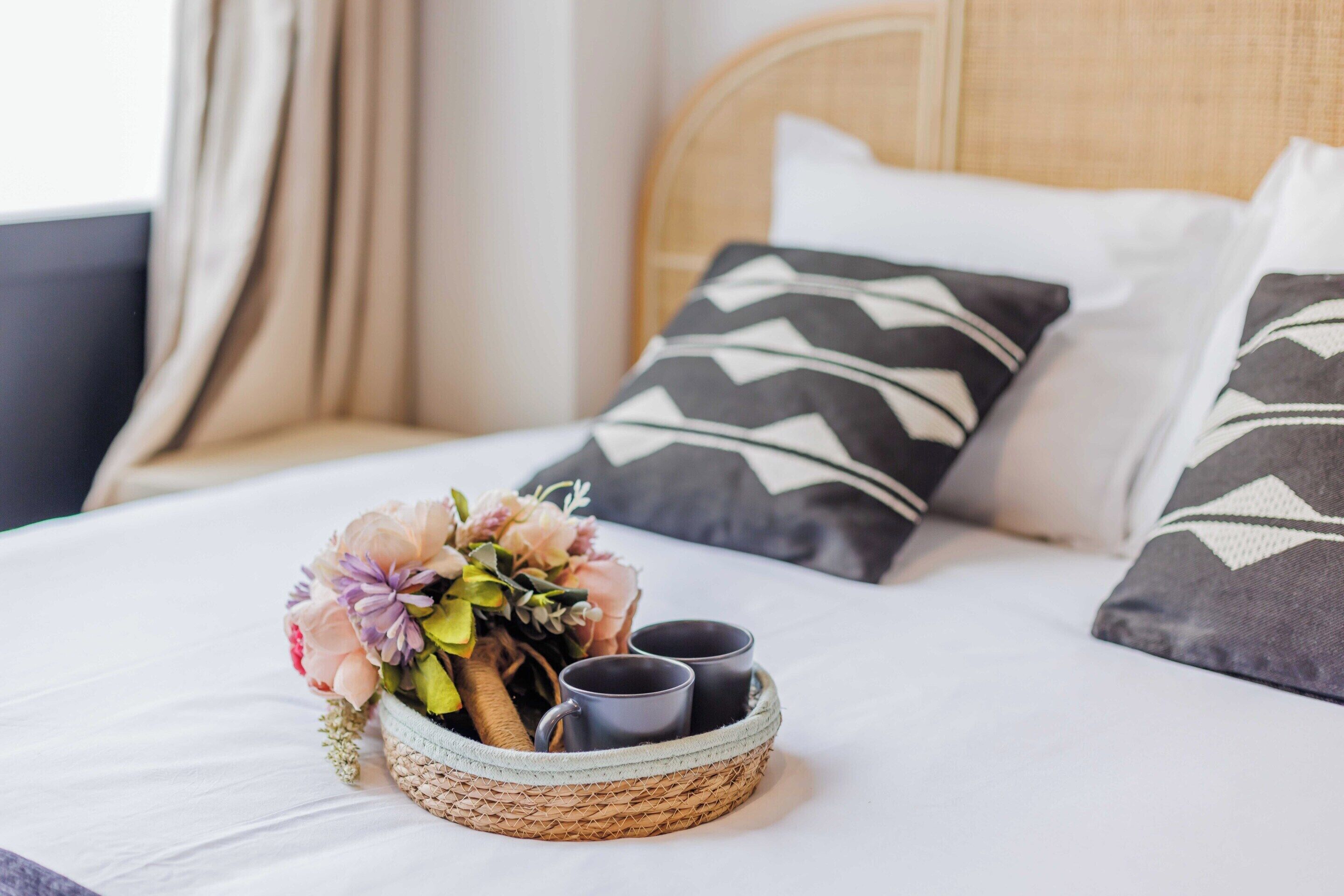 A neatly made bed with a woven basket containing a bouquet of flowers and two mugs, set against a backdrop of patterned pillows and a headboard.