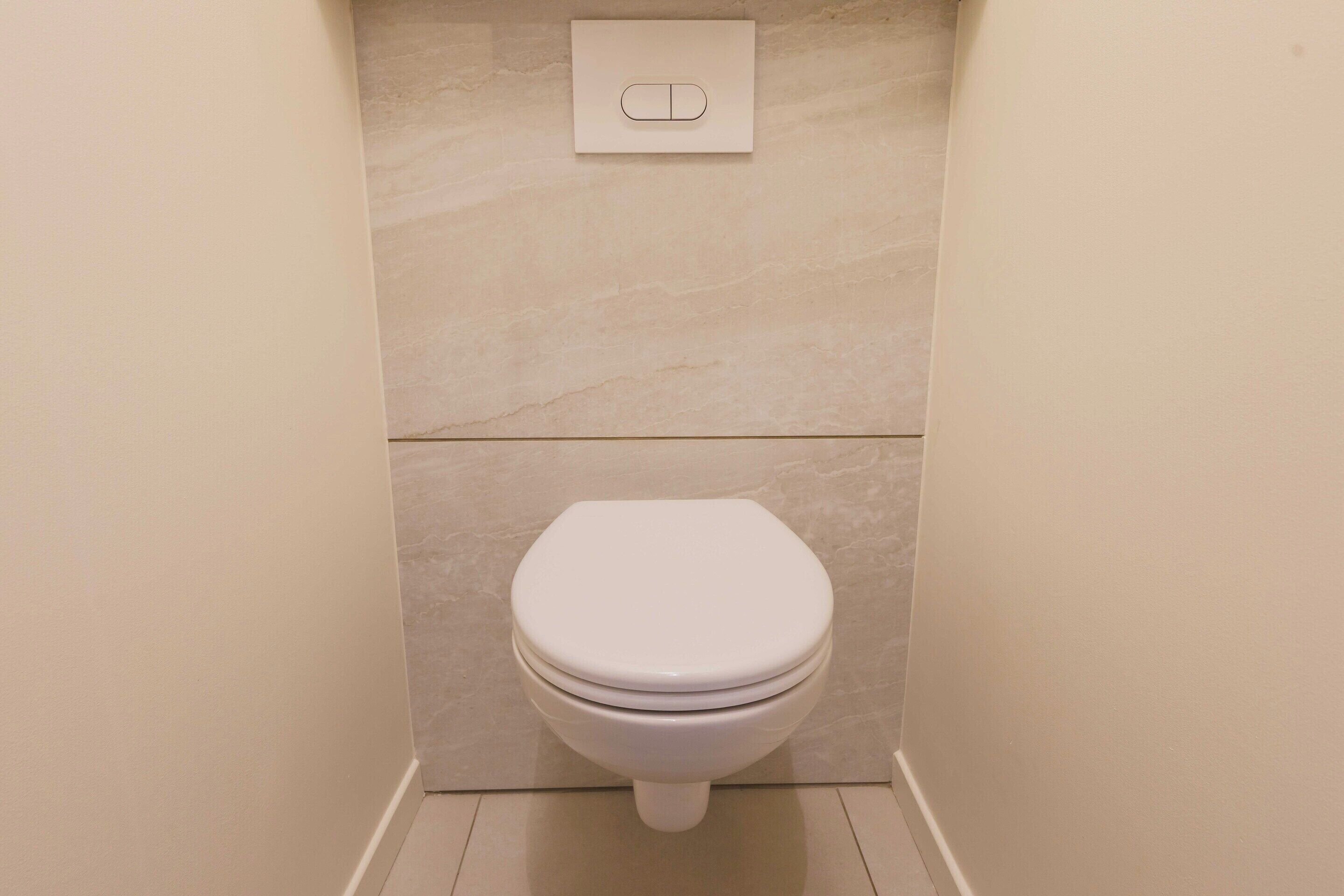 Un bagno piccolo e moderno, con il water, un pulsante a muro per lo sciacquone e pavimento piastrellato.