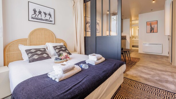 A hotel room with a bed, towels, a headboard, a wall with a picture, a door, and a hallway leading to a bathroom. - Wonderful Apartment 1br4p (Paris)