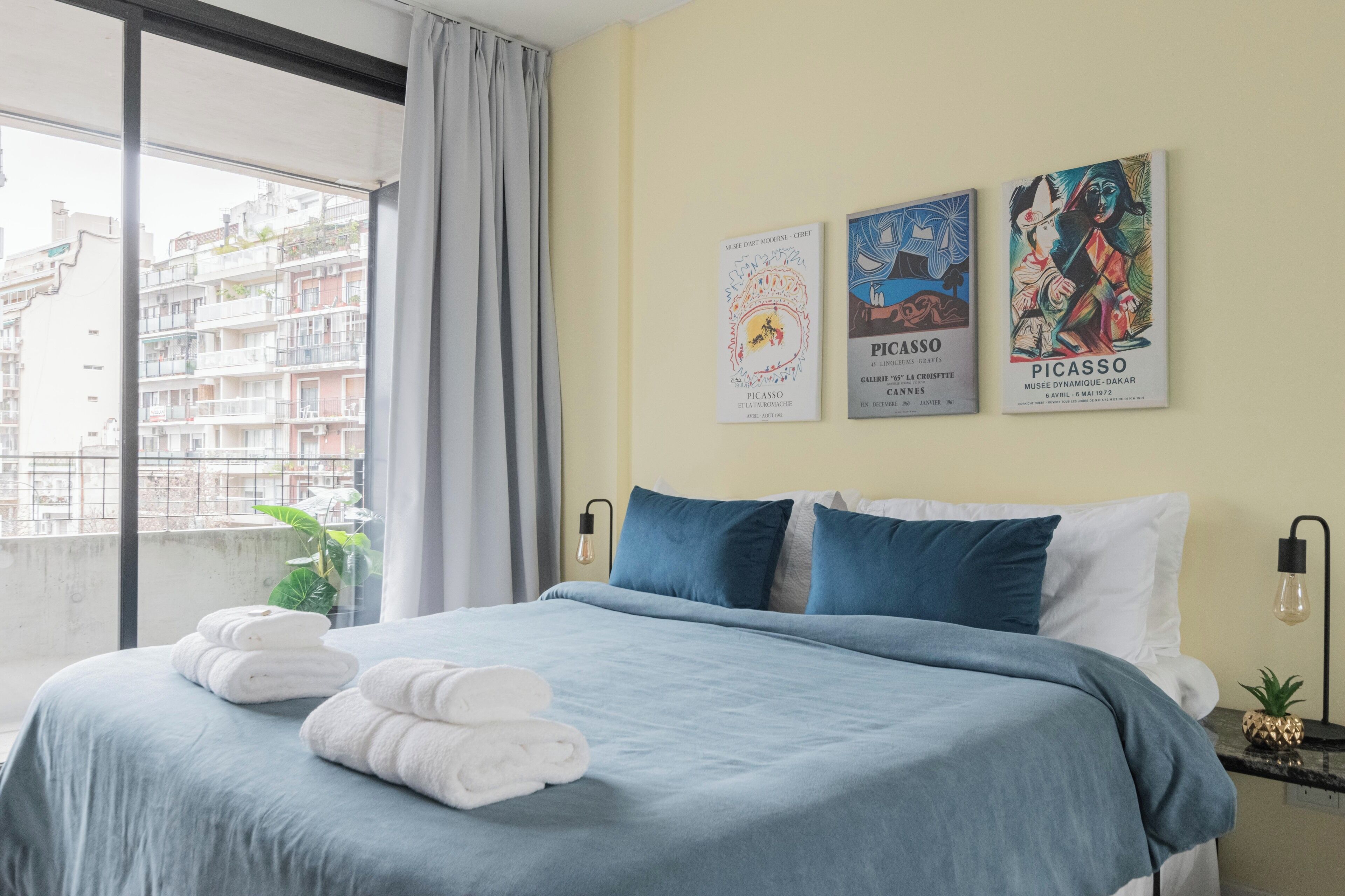 A hotel room with a large bed, a view of the city through a window, and two posters on the wall.
