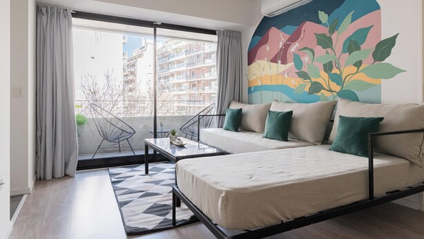 A modern bedroom with a sofa bed, a small table, and a chair. There is a large window with curtains, a colorful mural on the wall, and a view of buildings outside. - MOT ARHAUS (Buenos Aires)