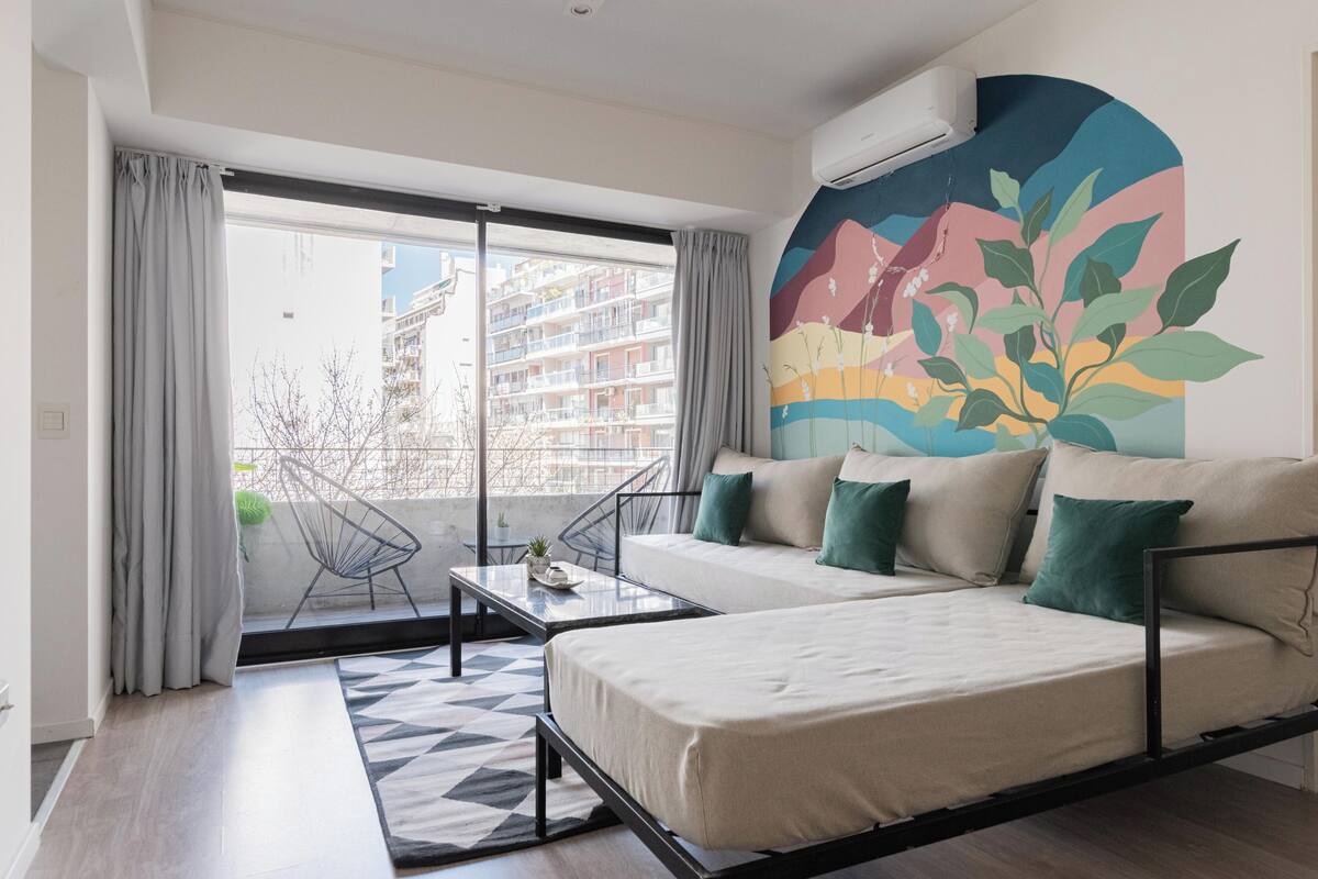 Un dormitorio moderno con sofá cama, una mesita y una silla. Hay una ventana grande con cortinas, un mural colorido en la pared y vistas de edificios.