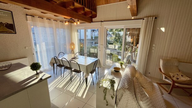 Un espace repas comprenant une table et des chaises, un canapé et un fauteuil, situé dans une pièce dotée de grandes fenêtres et offrant une vue sur l’extérieur.