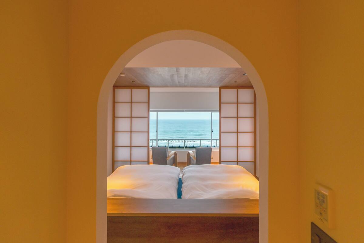 A bedroom with a view of the sea through a large window, a bed with white linens, and a wooden headboard.