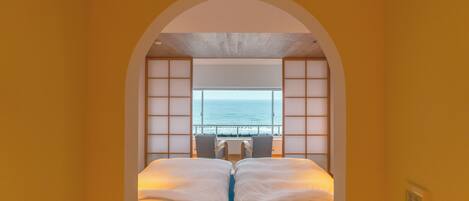 Une chambre avec vue sur la mer, grâce à une grande fenêtre, un lit avec du linge de lit blanc et une tête de lit en bois.