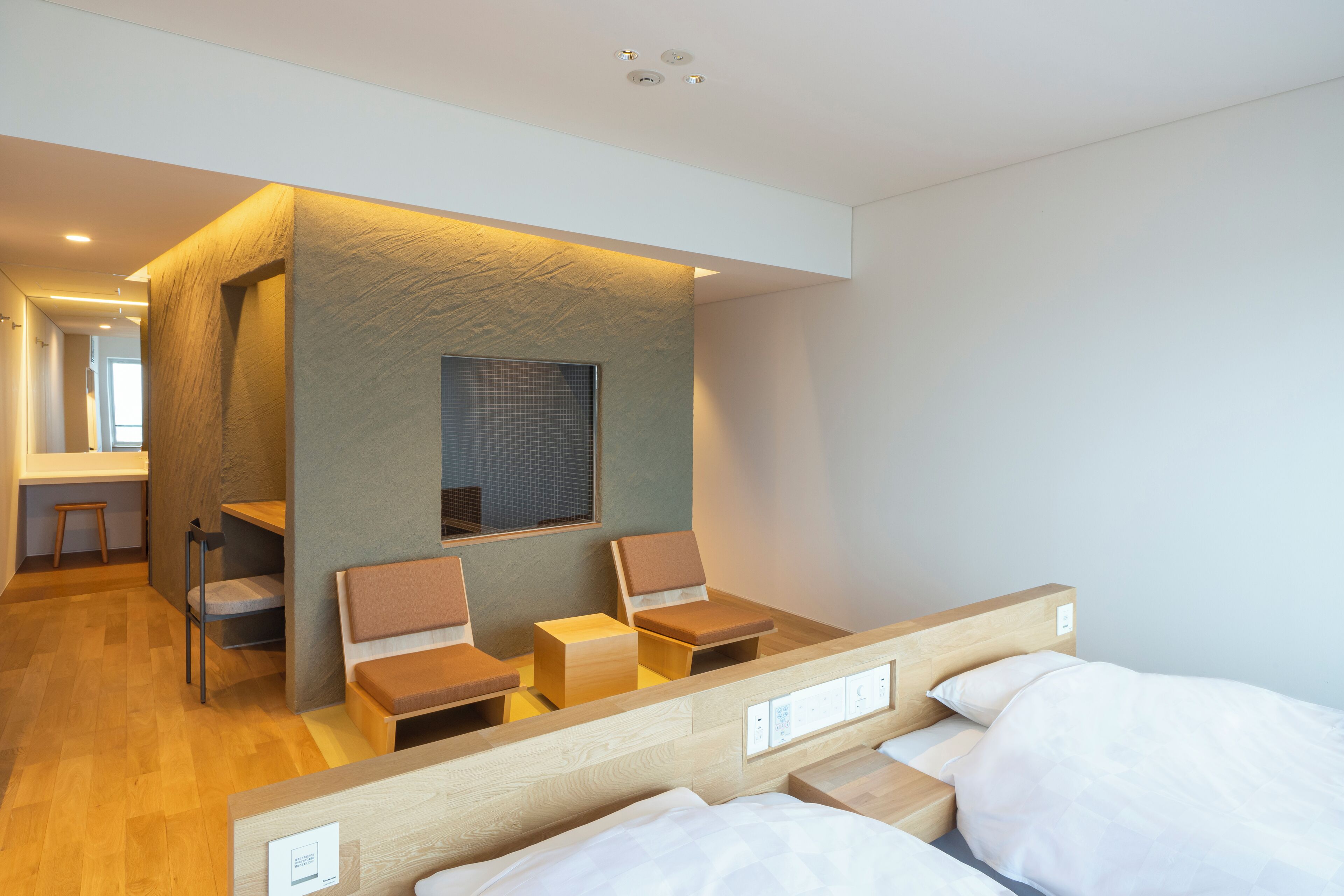 Une chambre d’hôtel moderne avec une tête de lit en bois, un petit coin salon avec des chaises et une table, et une grande fenêtre.