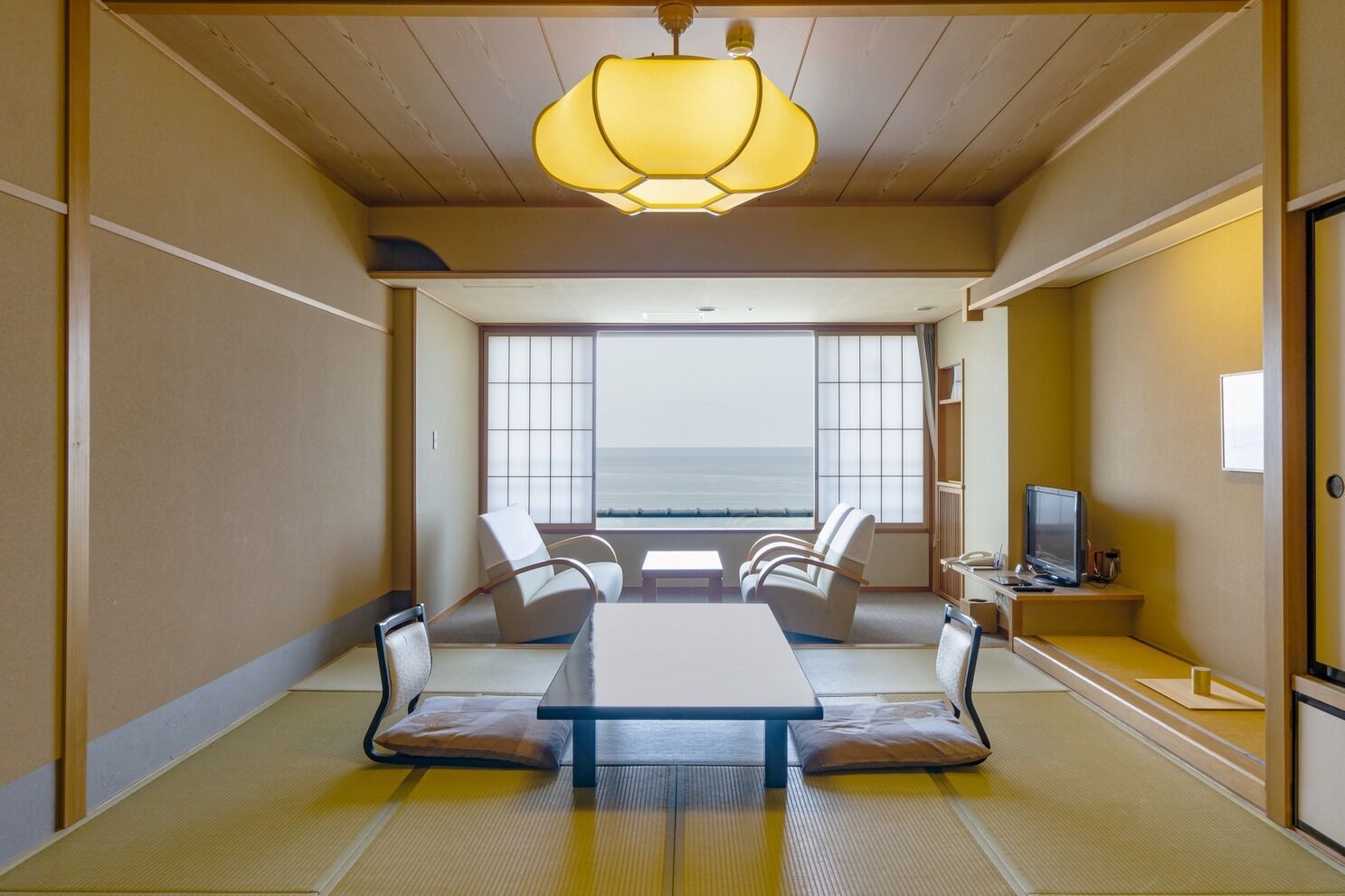 A room with tatami flooring, a low table, two chairs, a television, and a view of the sea.