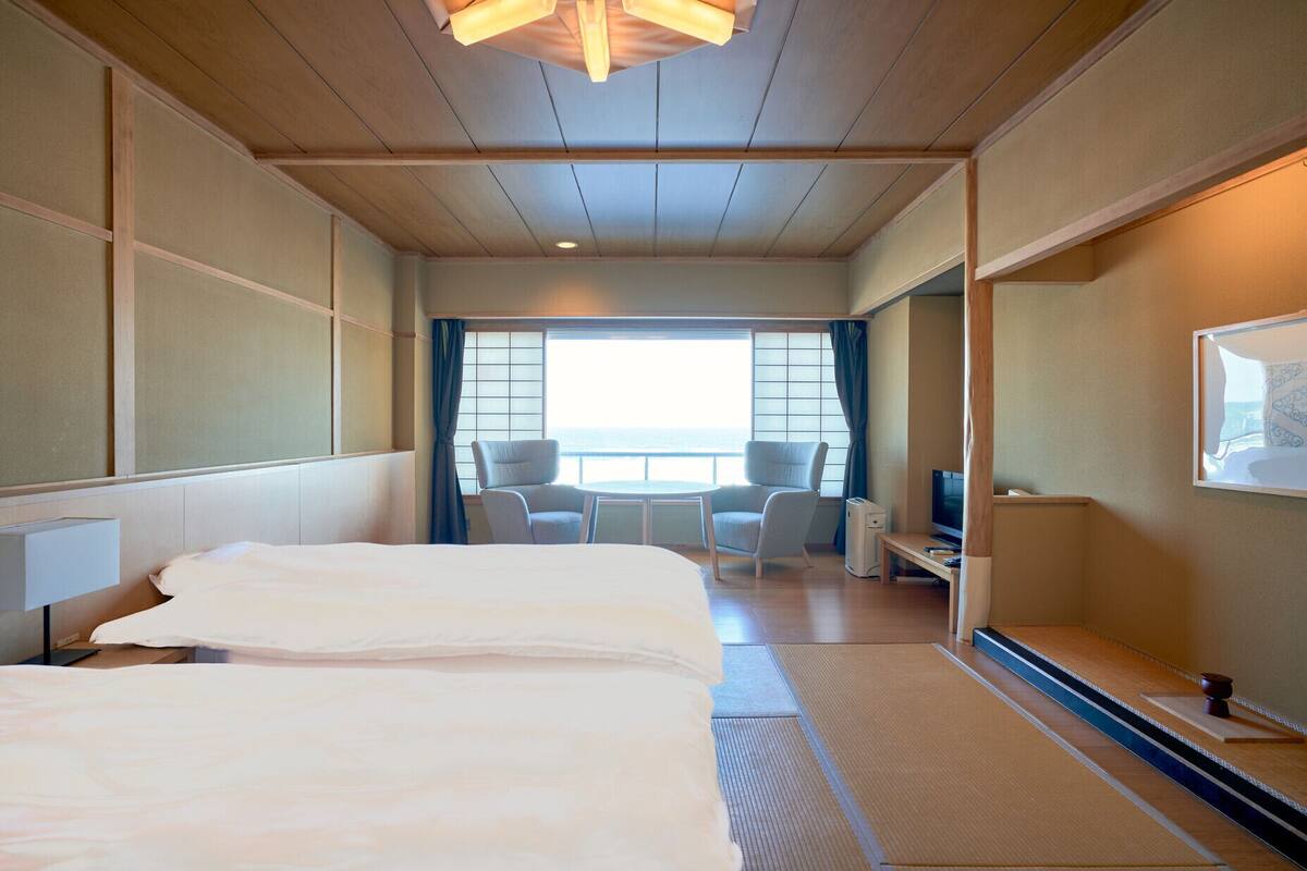 A hotel room with a bed, two chairs, a desk with a TV, and a large window with a view.