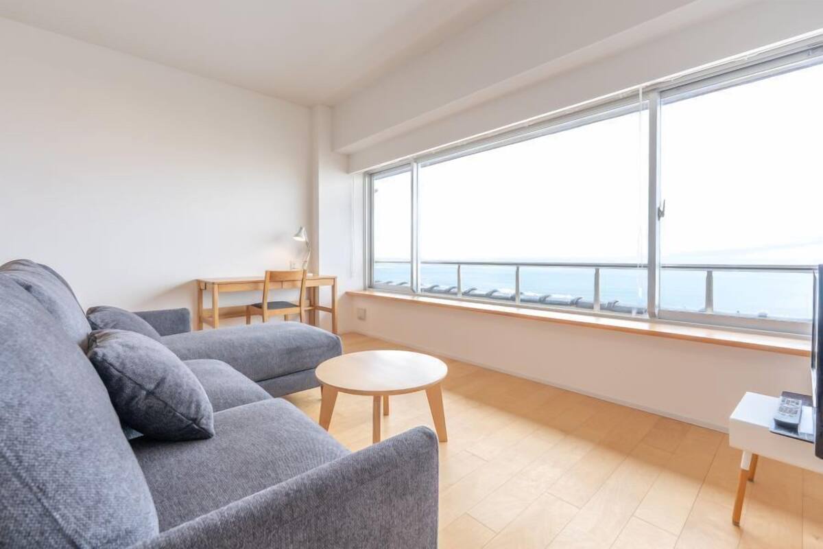 A modern living room with a grey sofa, a small round table, and a view of the sea through large windows.