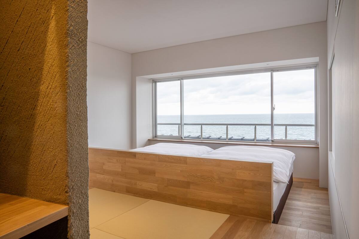 A bedroom with a large window overlooking the sea, a wooden bed, and a tatami mat floor.