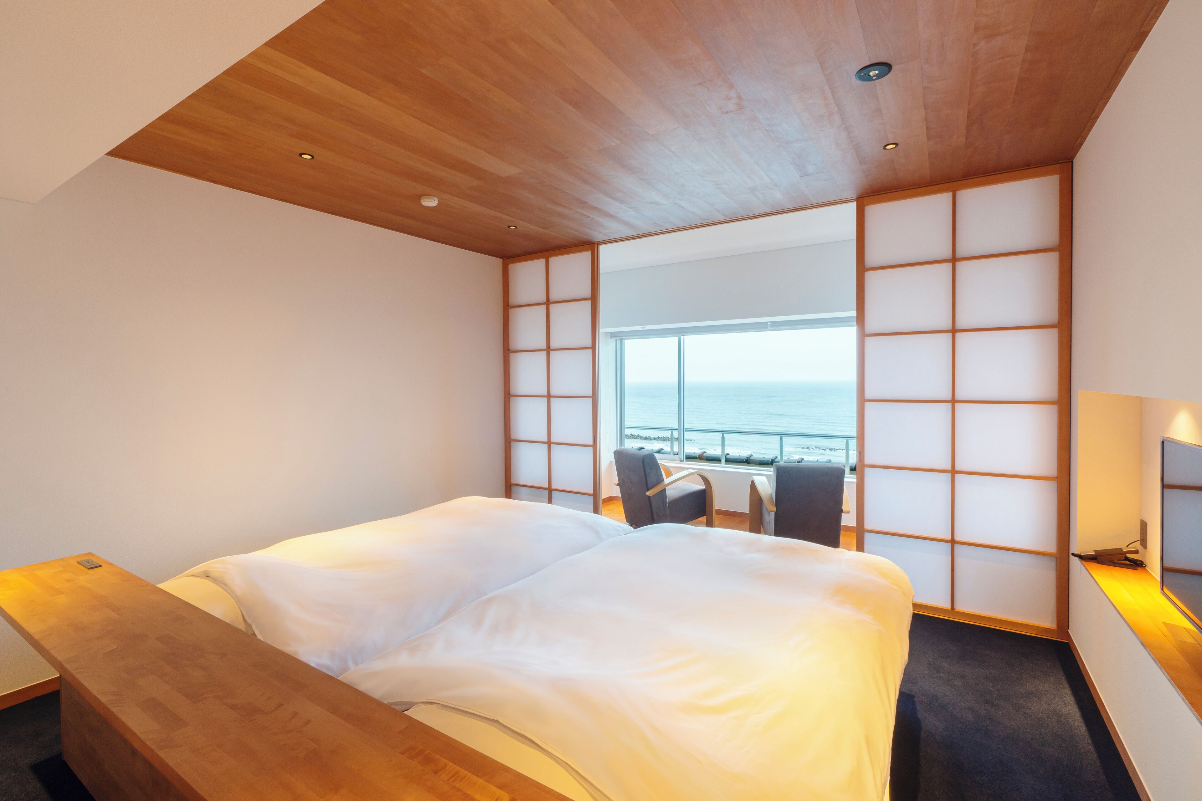 A modern hotel room with a wooden ceiling, a large bed, a seating area with a view of the sea, and a traditional Japanese sliding door.