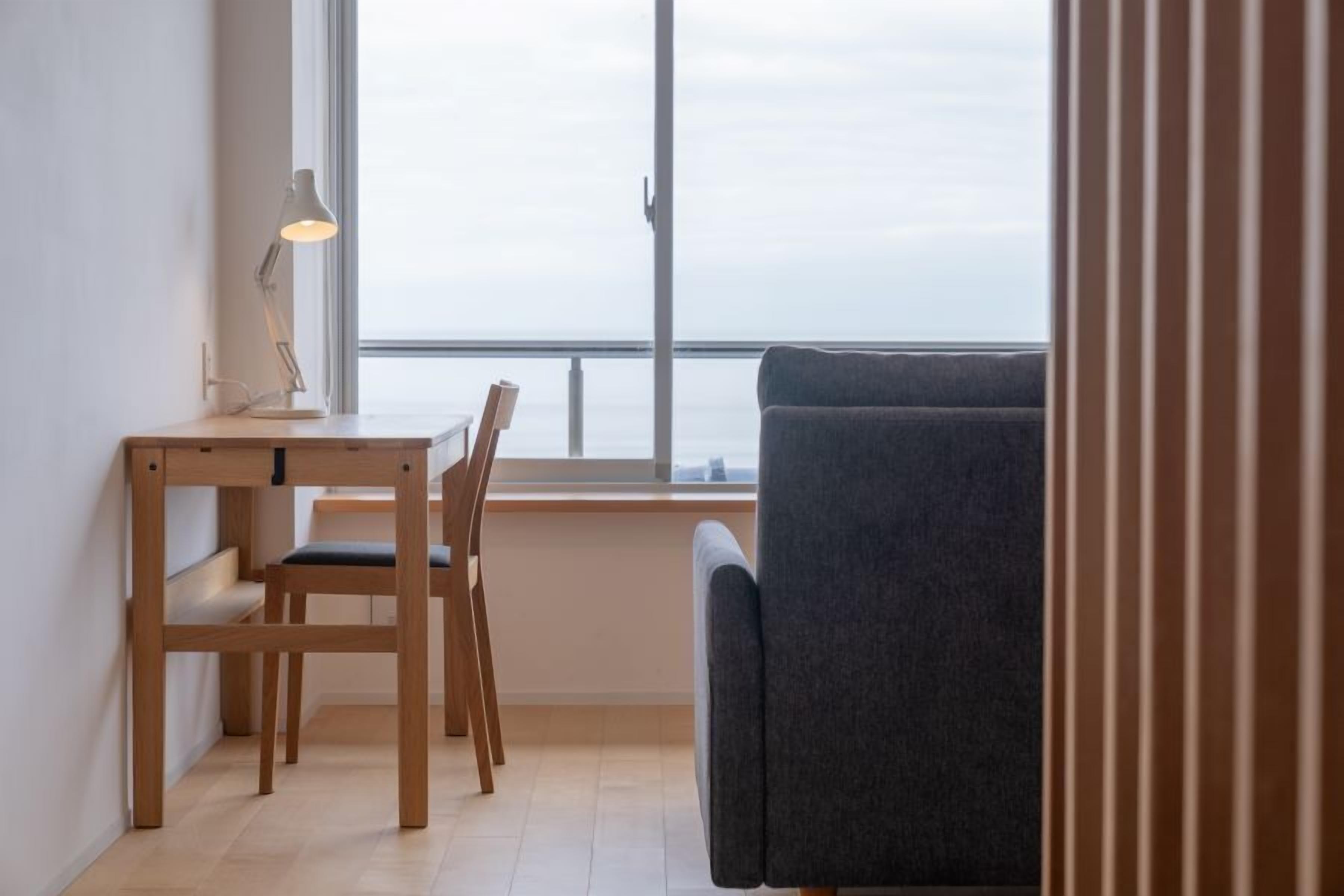 A room with a wooden desk, two chairs, a grey sofa, and a large window overlooking a body of water.