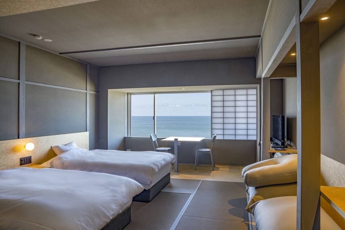 A hotel room with two beds, a desk, a chair, a TV, and a large window with a view of the sea.