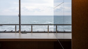 Vista da una stanza con una grande finestra che si affaccia sul mare, un corrimano in metallo e un pavimento in cemento.
