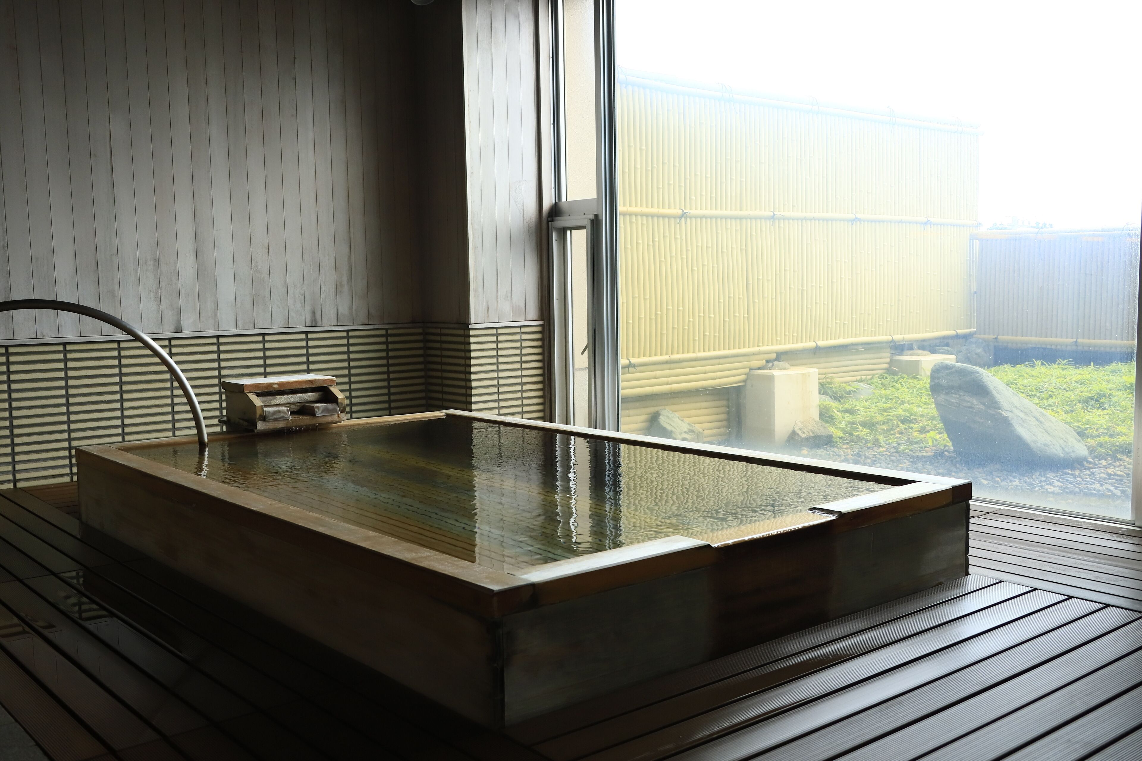 Uma onsen japonesa tradicional com uma banheira de madeira, piso de madeira e vista para um jardim através de uma grande janela.
