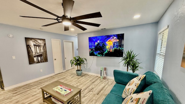 A living room with a ceiling fan, a mounted TV displaying a nature scene, a green sofa with patterned cushions, a coffee table with board games, and a door with a framed picture.