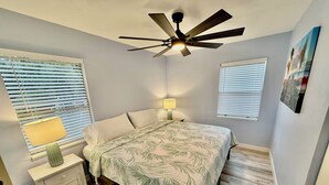 A bedroom with a bed, a nightstand, a lamp, a ceiling fan, and two windows with blinds.