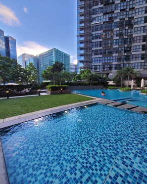 A swimming pool with tiled flooring, surrounded by greenery and a cityscape in the background. - Modern Stylish 1BR Condo Uptown BGC w/ View - 22u (Taguig)