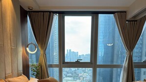 A hotel room with a large bed, beige bedding, and a view of a cityscape through floor-to-ceiling windows.