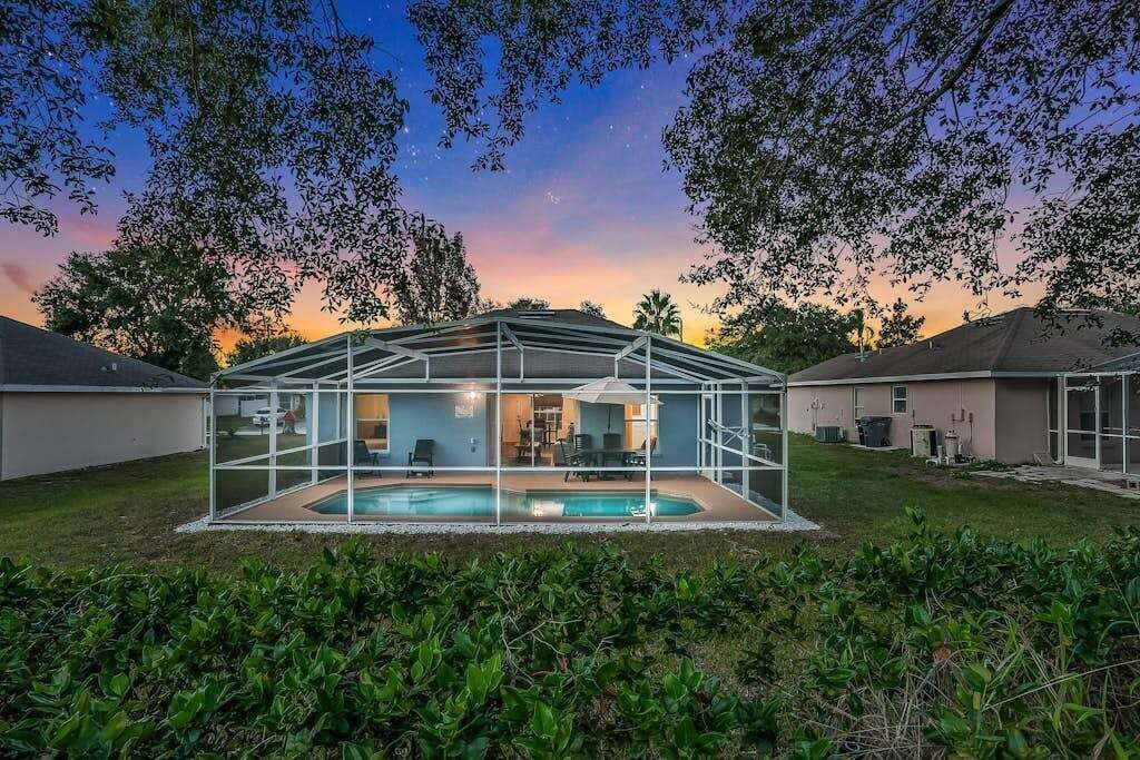 Un espace piscine entièrement vitré, offrant une vue sur le ciel au coucher du soleil, entouré de verdure et bordé de maisons.