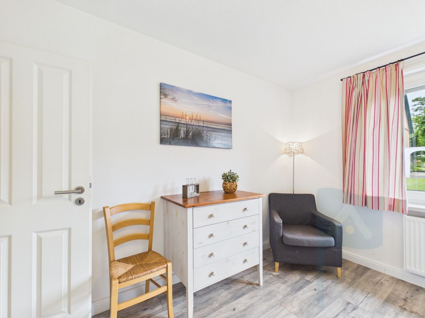 A room with a wooden chair, a white dresser with drawers, a gray armchair, a lamp, and a painting of a sunset.