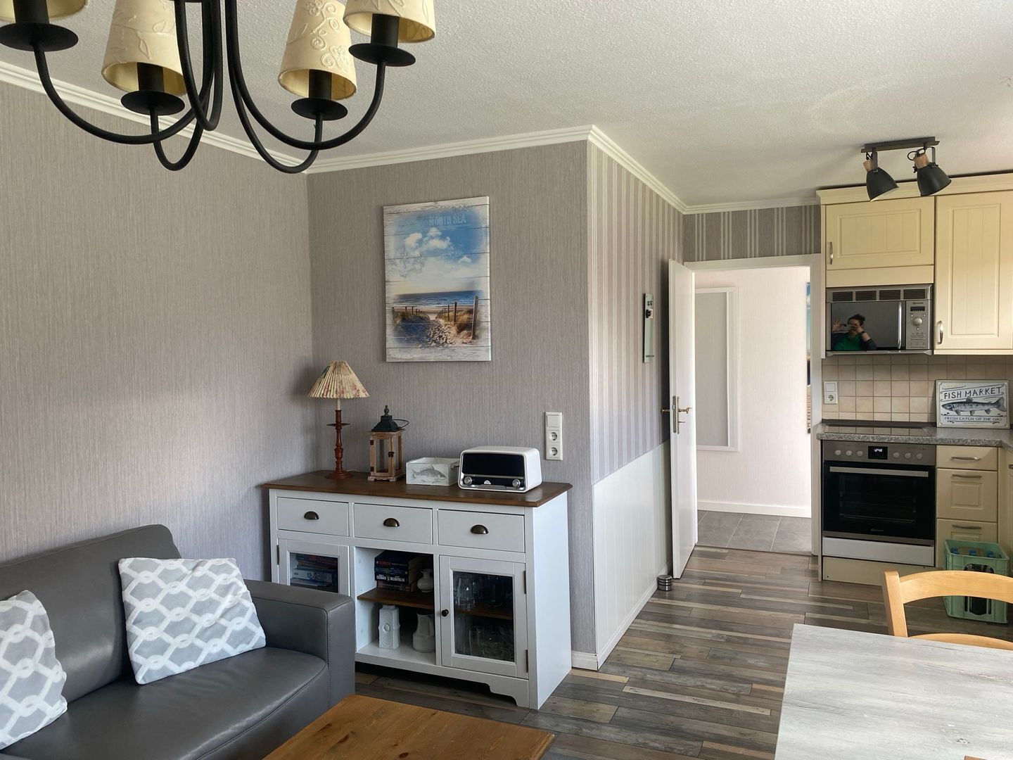 Une cuisine moderne avec des armoires blanches, un four à micro-ondes et une cuisinière. On y trouve une table à manger avec des chaises, un canapé avec des coussins à motifs et un lustre. Un tableau représentant une scène de plage est accroché au mur.